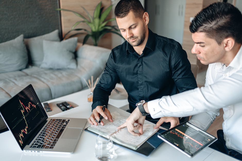 Two businessmen collaborating on stock market analysis with charts on laptops and tablets in a modern office.